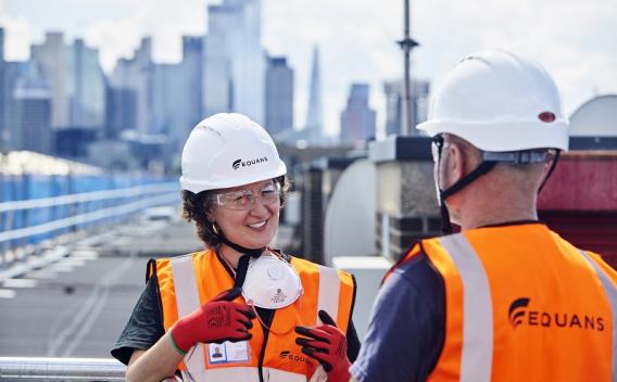 Two Equans employees stood on a rooftop wearing hard hats with chin straps
