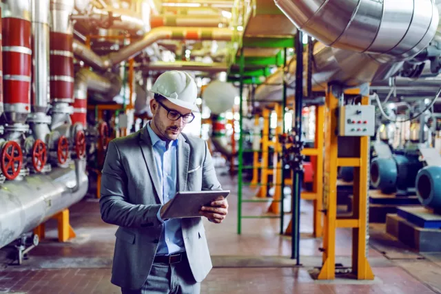 Stock image showing man in business suit wearing a hard hat, looking at an ipad in a plant room